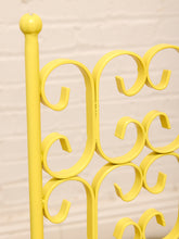 Load image into Gallery viewer, A close-up of a bright yellow metal garden stake or decorative gate frame with ornate scrollwork, set against a light beige brick wall background.
