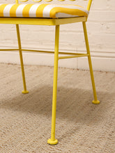 Load image into Gallery viewer, A close-up of a yellow metal chair with a yellow and white striped cushion, shown on a beige woven rug against a white brick wall.
