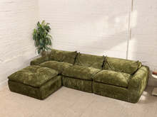 Load image into Gallery viewer, An olive green velvet sectional sofa with a matching ottoman, set in a room with a white brick wall and a potted plant.
