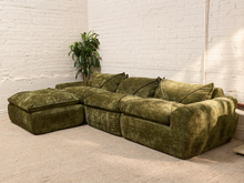 Load image into Gallery viewer, An olive green velvet sectional sofa with a chaise ottoman, set in a room with white brick walls and a beige carpet.
