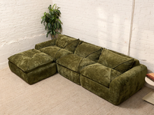 Load image into Gallery viewer, An olive green velvet sectional sofa with a matching ottoman, set in a room with a white brick wall and beige carpet.
