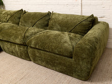 Load image into Gallery viewer, An olive green velvet sectional sofa with matching back cushions, shown in a room setting on a textured beige rug against a white brick wall.
