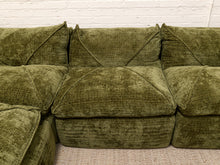 Load image into Gallery viewer, An olive green velvet sectional sofa with thick, oversized cushions, shown against a white brick wall and light beige carpet.
