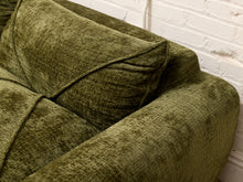 Load image into Gallery viewer, Close-up of an olive green textured fabric sofa cushion and armrest against a white painted brick wall.
