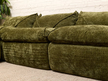Load image into Gallery viewer, An olive green velvet sectional sofa with plush cushions, set against a white brick wall and beige carpet.
