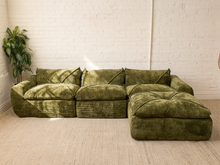 Load image into Gallery viewer, An olive green velvet sectional sofa with a matching ottoman, set in a minimalist room with a white brick wall and light beige carpet.
