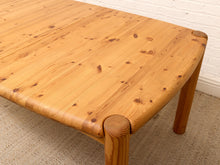Load image into Gallery viewer, A close-up of a light natural pine wooden table with rounded corners and thick legs, set on a textured beige rug against a white brick wall.
