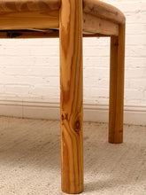 Load image into Gallery viewer, Close-up of a wooden table leg with a warm honey finish and visible wood grain, standing on a beige carpet against a white brick wall.
