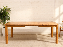 Load image into Gallery viewer, A rectangular wooden dining table with a natural bamboo top and rounded legs, set against a white brick wall with a potted plant on the left.
