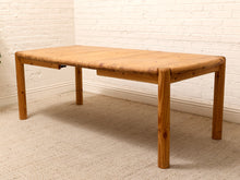 Load image into Gallery viewer, A rectangular wooden table with a light natural wood top and legs, positioned in a room with a white brick wall and beige carpet.
