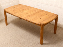 Load image into Gallery viewer, A rectangular wooden coffee table with a light natural pine finish, featuring rounded corners and thick cylindrical legs, set against a white brick wall and textured beige carpet.
