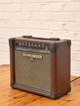 Load image into Gallery viewer, A vintage-style Behringer UL10000A tube amplifier guitar amp with a brown wood-grain finish, black controls, and a beige grille, sitting on a wooden table against a white brick wall.

