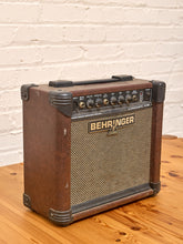 Load image into Gallery viewer, A vintage-style Behringer guitar amplifier with a brown wood-grain finish, black corner protectors, and a beige mesh grille, sitting on a wooden table against a white brick wall.
