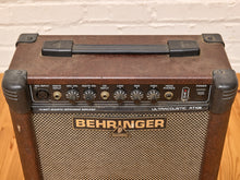 Load image into Gallery viewer, A vintage Behringer AT1020 acoustic guitar amplifier with a brown textured front, black controls, and a beige mesh grille, set against a white brick wall and wooden floor.
