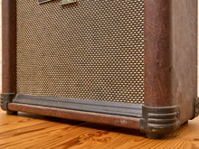 Load image into Gallery viewer, A close-up shot of a vintage-style guitar amplifier showing its brown textured grille cloth, dark metal corner protectors, and a weathered brown wooden cabinet resting on a wooden floor.
