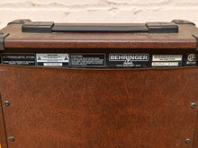 Load image into Gallery viewer, Close-up of the front of a Behringer UA-1000T acoustic guitar amplifier, showing the brown textured front panel and top handle against a white brick wall background.
