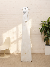 Load image into Gallery viewer, A tall, narrow white wooden wall art piece shaped like a stylized animal head with ears and a short tail, displayed against a white brick wall in a styled room with a chair and potted plant.
