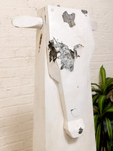 Load image into Gallery viewer, A rustic white wooden wall-mounted sculpture shaped like a cow head, with peeling paint and a small green plant visible against a white brick wall.
