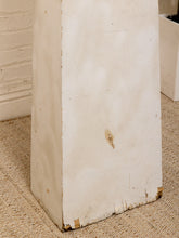 Load image into Gallery viewer, A close-up of a distressed, off-white painted wooden pedestal or column base on a textured beige carpet, with a white brick wall in the background.
