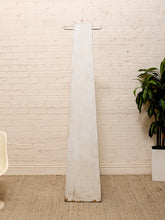 Load image into Gallery viewer, A tall, narrow rustic white wooden sculpture with a weathered finish stands against a white brick wall in a styled room setting with a potted plant and a chair partially visible.
