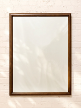 Load image into Gallery viewer, A tall rectangular mirror with a medium brown wooden frame, mounted on a white painted brick wall with soft leaf shadows cast across it.
