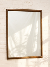 Load image into Gallery viewer, A rectangular wall mirror with a medium brown wooden frame, mounted on a white painted brick wall with soft palm leaf shadows cast across it.
