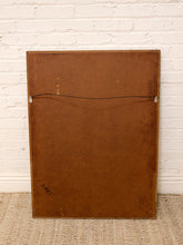 Load image into Gallery viewer, The back of a framed mirror, showing a brown cardboard backing with a hanging wire and small printed labels, leaning against a white brick wall on a woven rug.
