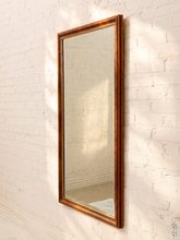 Load image into Gallery viewer, A tall, narrow rectangular wall mirror with a distressed bronze-brown wooden frame, mounted on a white painted brick wall.
