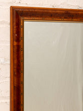 Load image into Gallery viewer, A close-up of a large wall mirror with a thick, polished reddish-brown wooden frame featuring brass nailhead trim, set against a white brick wall.
