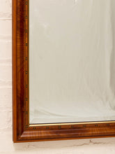Load image into Gallery viewer, A close-up of a large wall mirror with a richly carved warm brown wooden frame and brass-toned metal studs, mounted on a white painted brick wall.
