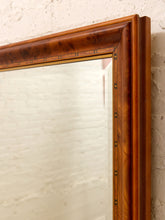 Load image into Gallery viewer, A close-up of a large rectangular mirror with an ornate warm brown wooden frame featuring brass corner hardware, set against a textured off-white brick wall.
