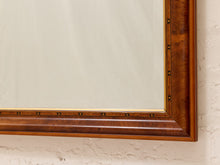 Load image into Gallery viewer, Close-up of a wooden wall mirror frame with a warm medium-brown wood grain and brass-toned metal accents, mounted on a textured off-white plaster wall.
