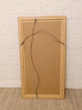 Load image into Gallery viewer, The back of a wooden picture frame with a light natural wood border, a brown cardboard backing, and a hanging wire with clips, leaning against a white brick wall on a woven rug.
