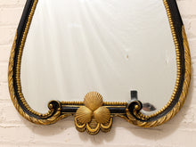 Load image into Gallery viewer, An ornate oval wall mirror with a black and gold decorative frame featuring shell and scroll motifs, mounted on a white brick wall.
