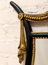 Load image into Gallery viewer, Close-up of an ornate vintage-style mirror frame with a glossy black background and intricate gilded gold detailing, including a decorative hanging ornament and beaded trim, set against a textured off-white brick wall.
