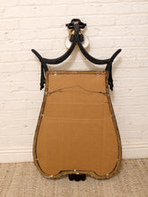 Load image into Gallery viewer, An antique-style wall mirror with a black metal scrollwork frame and a large tan cardboard backing, photographed against a white brick wall.
