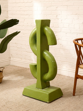 Load image into Gallery viewer, A sculptural, abstract green sculpture with a rectangular base and curved, loop-like upper structure, shown in a styled room corner with a white brick wall, beige carpet, a plant, and part of a wooden chair.
