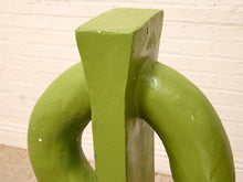 Load image into Gallery viewer, A close-up of a green abstract sculptural object with curved forms and a vertical rectangular section, photographed indoors against a white painted brick wall.
