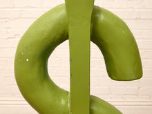 Load image into Gallery viewer, A close-up of a green dollar sign sculpture or decorative object painted in a matte olive green, set against a white painted brick wall.

