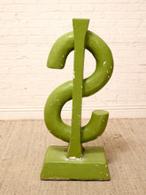 Load image into Gallery viewer, A green dollar sign sculpture on a square base, set against a white brick wall and beige carpet.
