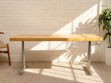 Load image into Gallery viewer, A modern writing desk with a light natural wood top and silver metal legs, set against a white brick wall with warm sunlight and leaf shadows.
