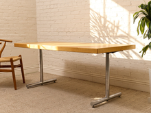 Load image into Gallery viewer, A modern rectangular desk with a light natural wood top and a silver metal T-style pedestal base, set in a bright room with a white brick wall, beige carpet, and a wooden chair partially visible on the left.

