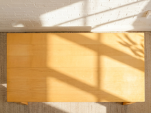 Load image into Gallery viewer, A top-down view of a light natural wood rectangular table on a woven rug, with a white brick wall and strong diagonal window shadows cast across the tabletop.

