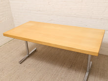 Load image into Gallery viewer, A light natural wood rectangular table with a silver metal T-style base, shown in a room setting with a white brick wall and beige carpet.
