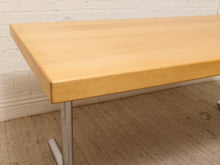 Load image into Gallery viewer, A light natural wood writing desk with a silver metal pedestal leg, shown in a room setting with a white brick wall and carpeted floor.
