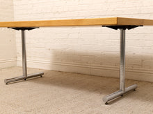 Load image into Gallery viewer, A rectangular table with a light natural wood top and silver metal pedestal legs, shown in a room with a white brick wall and beige carpet.
