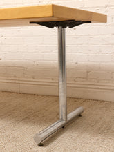 Load image into Gallery viewer, A close-up of a table leg assembly with a light wood top, black support brackets, a vertical brushed metal tube, and a horizontal base on a beige carpet against a white brick wall.
