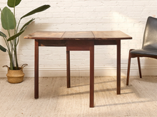 Load image into Gallery viewer, A dark brown wooden extendable table with a rectangular top and four legs, set against a white brick wall with a potted plant on the left and a chair on the right.
