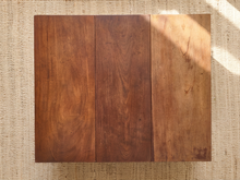 Load image into Gallery viewer, A rectangular wooden board or tabletop made of three vertical panels with warm medium-to-dark brown wood grain, photographed from above on a beige woven rug with soft sunlight and shadows.
