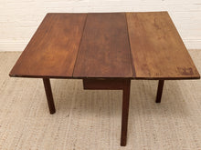 Load image into Gallery viewer, A three-panel wooden drop-leaf table with a warm medium-brown finish, shown on a textured beige rug against a white brick wall.

